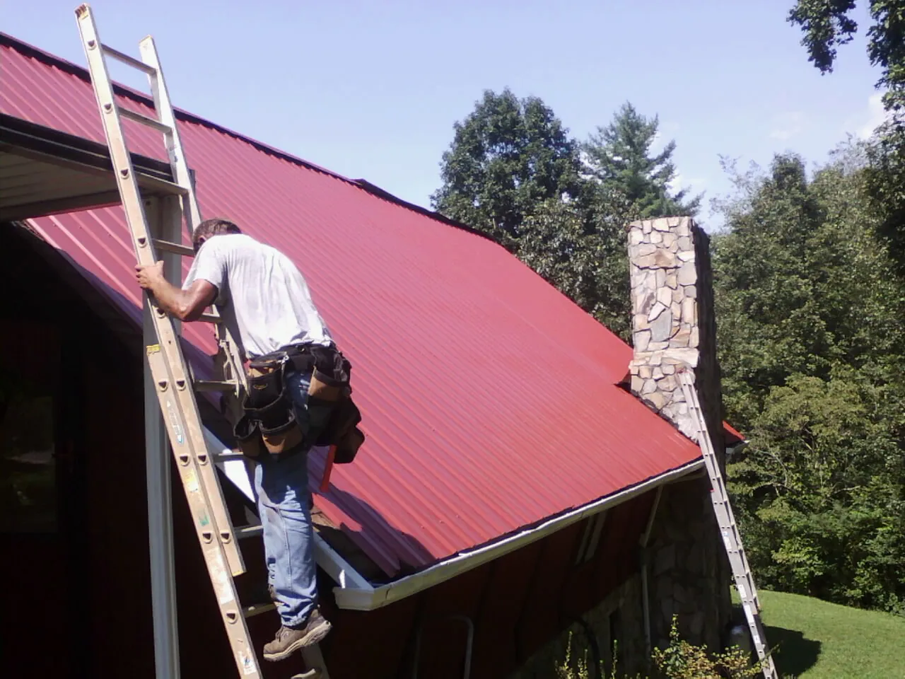 Roofer on a ladder beside a red metal roof and stone chimney — Soffit & Fascia Repair by Roofing Brothers in Teachey